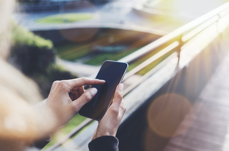 Hipster texting message on smartphone or technology, mock up of blank screen. Tourist girl using cellphone on bridge background close. Female hands holding gadget on blurred backdrop. Mockup copy space for text message or design flareの写真素材