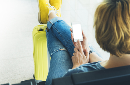 Young hipster girl at airport and put feet in yellow boot on suitcase waiting air flight, female hands holding smart phone in terminal departure lounge gate, traveler trip concept, mockup of blank screen sellphoneの写真素材