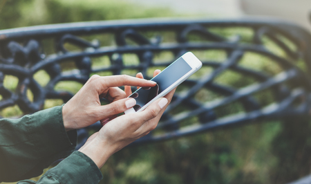 Hipster texting message on smartphone or technology, mock up of blank screen. Tourist girl using cellphone on forged steel bench background close. Female hands holding device. Mockup copy space for text messageの写真素材