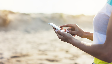 Hipster girl hold on smart phone gadget in sand coastline, mock up blank screen. Traveler using in female hand mobile on background beach seascape horizon. Tourist look on blue sun ocean, summer lifestyleの写真素材