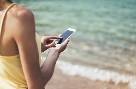 Hipster girl hold on smart phone gadget in sand coastline, mock up blank screen. Traveler using in female hand mobile on background beach seascape horizon. Tourist look on blue sun ocean, summer lifestyleの写真素材
