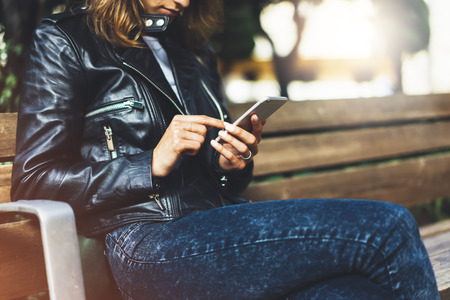 Girl in leather jacket holding smart phone on bench background sun atmospheric city, hipster using in female hands and texting mobile, street lifestyle, tourist planing route in summer conceptの写真素材