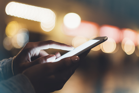 Girl pointing finger on screen smartphone on background illumination bokeh color light in night atmospheric city, hipster using in female hands and texting mobile phone, mockup glitter street, content lifestyleの写真素材
