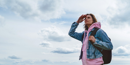 tourist traveler with black backpack on background mountain, hiker looks up at blue sky clouds, girl enjoying nature panoramic landscape in trip, relax holiday mockup concept in trekking tripの写真素材