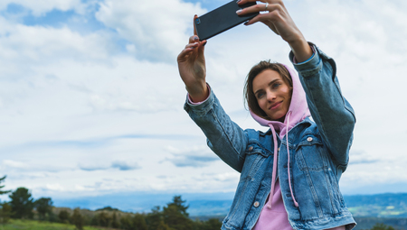 tourist girl on background blue sky clouds taking photo selfie on mobile smart phone, person looking on camera gadget technology, blogger using content online wifi internet lifestyle conceptの写真素材