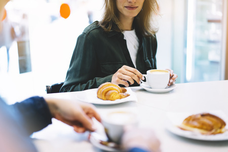 Young woman holding in female hands croissant and drink hot aroma coffee or tea in breakfast time, hipster traveler smile girl communicate in the morning sun day, holiday relax white cafe with friendsの写真素材
