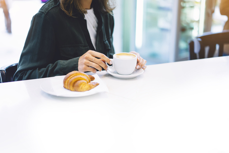 Young woman holding in female hands croissant and drink hot aroma coffee or tea in breakfast time, hipster traveler smile girl communicate in the morning sun day, holiday relax white cafe with friendsの写真素材