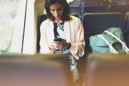 Enjoying travel. Young pretty woman traveling by train sitting near the window using smartphone and looking map. Tourist texting online message and plan route of railway, railroad transport conceptの写真素材