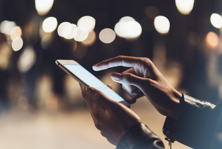 Girl pointing finger on screen smartphone on background illumination bokeh color light in night atmospheric city, hipster using in hands and texting mobile phone, mockup glitter street, content lifestyleの写真素材
