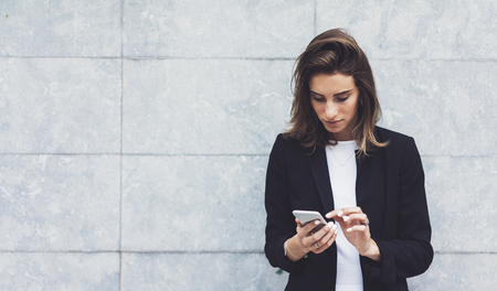 Portrait young businesswomen in black suit using smartphone isolated on background concrete gray wall mockup, pretty hipster manager holding mobile gadget, girl smiletexting message, connectの写真素材