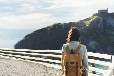 Hipster young girl with backpack enjoying sunset on seascape on peak mountain. Tourist traveler on background valley landscape view mockup. Hiker looking ocean and house on hill island in trip holidayの写真素材