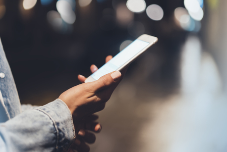 Girl pointing finger on screen smartphone on background illumination bokeh color light in night atmospheric city, hipster using in female hands and texting mobile phone, mockup glitter street, content lifestyleの写真素材
