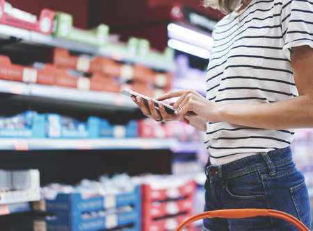 Young woman shopping healthy food in supermarket blur background. Close up view girl buy products using smartphone in store. Hipster at grocery using smartphone. Person comparing the price of produce of with her digital gadget at store.の写真素材