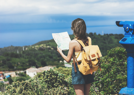 Hipster young girl with bright backpack looking at a map and poining hand the travel plan. View from the back of the tourist traveler on background mountain, sea, coin operated binoculars. Mock up for text messageの写真素材