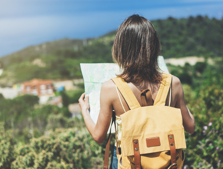 Hipster young girl with bright backpack looking at a map and poining hand the travel plan. View from the back of the tourist traveler on background mountain, sea. Mock up for text messageの写真素材