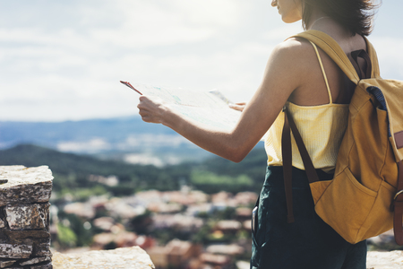Hipster young girl with bright backpack looking at a map and poining hand the travel plan. View from the back of the tourist traveler on background mountain, sea, coin operated binoculars. Mock up for text messageの写真素材
