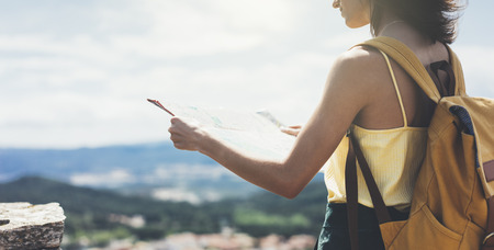 Hipster young girl with bright backpack looking at a map and poining hand the travel plan. View from the back of the tourist traveler on background mountain, sea, coin operated binoculars. Mock up for text messageの写真素材