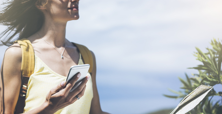 Hipster young girl with bright backpack looking at phone and map. Said view tourist traveler on background mountain, blue sea. Mock up for text message. Female hands using smartphone, holding gadgetの写真素材
