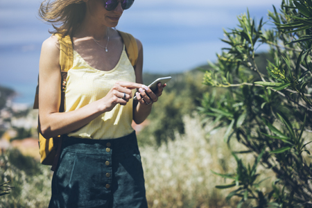 Hipster young girl with bright backpack looking at phone and map. Said view tourist traveler on background mountain, blue sea. Mock up for text message. Female hands using smartphone, holding gadgetの写真素材