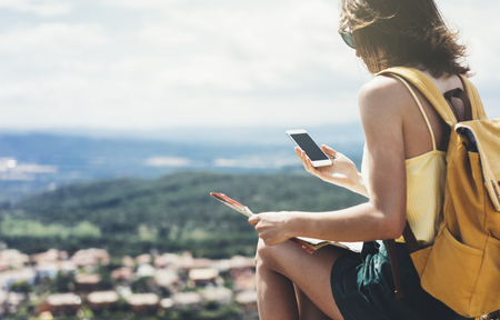Hipster young girl with bright backpack looking at a map. View from the back of the tourist traveler on background mountain, sea. Mock up for text message. Female hands using smartphone, holding gadgetの写真素材