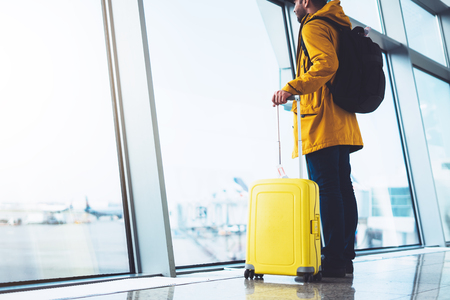 tourist with yellow suitcase backpack is standing at airport on background large window, traveler man waiting in departure lounge area hall of ââthe airport lobby terminal, vacation trip conceptの写真素材