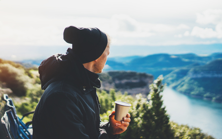 hipster tourist hold in hands mug of hot drink, lonely guy enjoy sun flare mountain in auto, traveler drink cup of tea on nature, vacation weekend concept on background of panoramic landscapeの写真素材