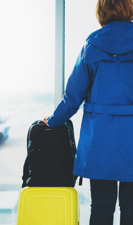 traveler tourist with yellow suitcase backpack is standing at airport on background large window, girl in bright jacket waiting in departure lounge area hall of â airport lobby terminal, vacation trip concept, empty space mockupの写真素材