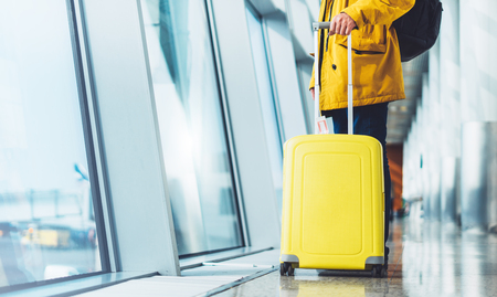 traveler tourist with yellow suitcase backpack at airport on background large window blue sky, man in bright jacket waiting in departure lounge area, hall of âairport lobby terminal, vacation trip concept, empty space mockupの写真素材