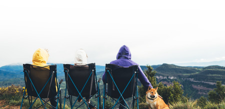 three friends sit in camping chairs on top of a mountain, travelers enjoy nature and cuddle with dog, tourists look into distance on background of panoramic landscape, weekend conceptの写真素材