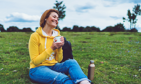 Happy smile girl holding in hands cup of hot tea on green grass in outdoors nature park, beautiful young woman hipster enjoy drinking cup of coffee, lifestyle relax recreation meditation conceptの写真素材