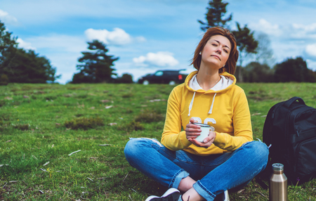 Happy girl with eyes closed holding in hands cup of hot tea on green grass in outdoors nature park, beautiful woman hipster enjoy drinking cup of coffee, lifestyle relax recreation meditation conceptの写真素材