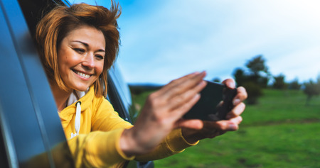 smile tourist girl in an open window of a auto car taking photo selfie on mobile smart phone, person looking on camera gadget technology, blogger using content online wifi internet conceptの写真素材