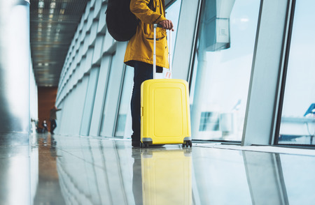 traveler in bright jacket with yellow suitcase backpack at airport on background large window blue sky, passenger waiting flight in departure lounge area, hall of âairport lobby terminal, vacation trip concept, empty space mockupの写真素材