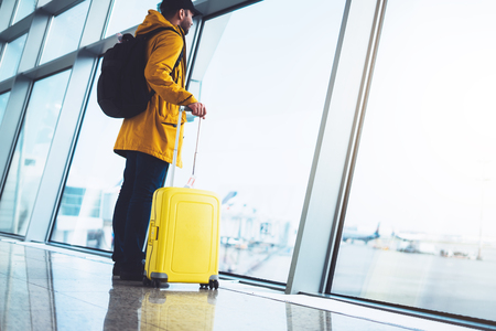tourist with yellow suitcase backpack is standing at airport on background large window, traveler person waiting in departure lounge area hall of ââthe airport lobby terminal, vacation trip conceptの写真素材