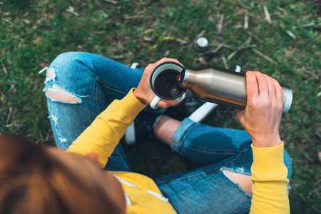 Top view girl holding in hands cup of hot tea on green grass in outdoors nature park, beautiful woman hipster enjoy drinking cup of coffee thermos, lifestyle relax recreation meditation conceptの写真素材