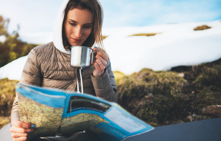 girl hold in hands cup of drinks, relax tourist look on map, people planning trip in snow mountain, hipster enjoy on background winter nature, journey landscape trip, lifestyle holiday conceptの写真素材