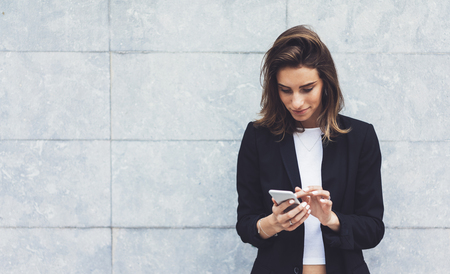Portrait young businesswomen in black suit using smartphone isolated on background concrete gray wall mockup, pretty hipster manager holding mobile gadget, girl smiletexting message, connectの写真素材