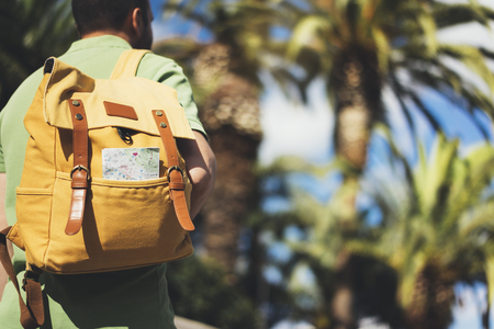 Hipster young man with backpack and map. View from a back tourist traveler planning route on background sun city and palm tree. Person visiting the sights Barcelona on backdrop summer street, mockupの写真素材
