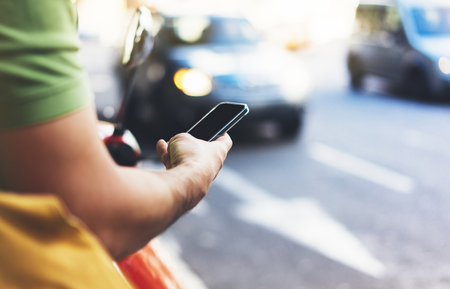Man pointing on screen smartphone on background cars, tourist hipster waiting using in hands mobile phone, traveler connect wifi internet and calls taxi, headlights auto on backdrop city street, mockupの写真素材