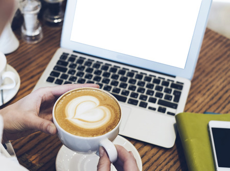 Woman looking on open laptop on wood table with empty blank screen monitor at home, person drink cup of coffee on background atmosphere cafe, female hands working on computer, mockup templatesの写真素材