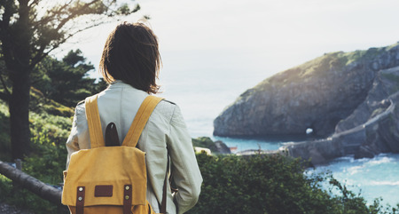 Hipster young girl with backpack enjoying sunset on seascape on peak mountain. Tourist traveler on background valley landscape view mockup. Hiker looking sunlight ocean in trip holiday in basque islandの写真素材