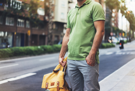 Hipster young man holding in hands backpack with map. Front view tourist traveler planning route and standing at the road on background sun summer street. Person hiker visiting the sights city , mockupの写真素材