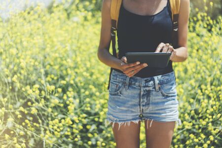 Hipster texting message on tablet or technology, mock up of blank screen. Girl using computer on yellow flowers background close. Female hands holding gadget on blurred backdrop. Mockup side view. Copy space for text message or designの写真素材