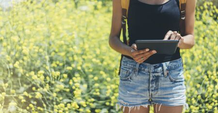 Hipster texting message on tablet or technology, mock up of blank screen. Girl using computer on yellow flowers background close. Female hands holding gadget on blurred backdrop. Mockup side view. Copy space for text message or designの写真素材