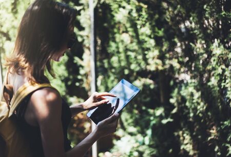 Hipster young girl with bright backpack and vintage camera using tablet or holding gadget, planning travel plan. View tourist traveler with sunglasses on background Parc Laberint Barcelona. Mock up for text messageの写真素材