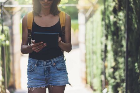 Hipster young girl with bright backpack and vintage camera using tablet or holding gadget, planning travel plan. View tourist traveler with sunglasses on background Parc Laberint Barcelona. Mock up for text messageの写真素材
