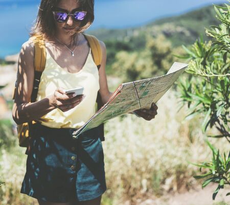 Hipster young girl with bright backpack looking at phone and map. Said view tourist traveler on background mountain, blue sea. Mock up for text message. Female hands using smartphone, holding gadgetの写真素材
