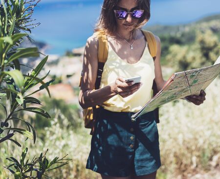 Hipster young girl with bright backpack looking at phone and map. Said view tourist traveler on background mountain, blue sea. Mock up for text message. Female hands using smartphone, holding gadgetの写真素材