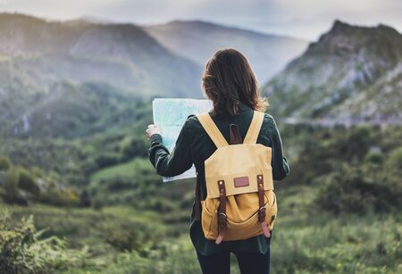 Hipster young girl with bright backpack enjoying sunset on peak of foggy mountain, looking a map and poining hand. Tourist traveler on background valley landscape panoramic view mockup, sunlight in trip in northern spain basque country, mock up for text.の写真素材