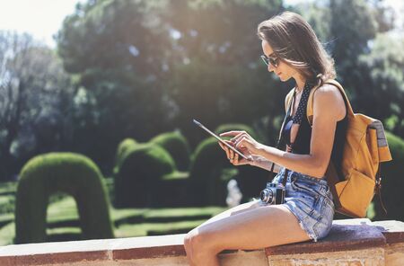 Hipster young girl with bright backpack and vintage camera using tablet or holding gadget, planning travel plan. View tourist traveler with sunglasses on background Parc Laberint Barcelona. Mock up for text messageの写真素材